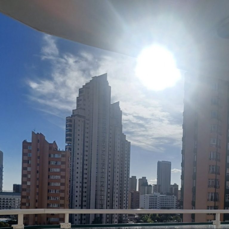 City skyline featuring tall buildings under a bright sun and blue sky.