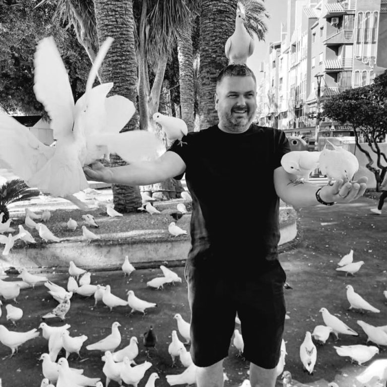 Man holding doves in a park surrounded by pigeons on the ground.