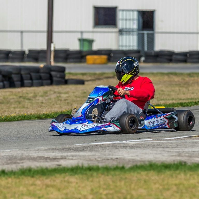 A go-kart racer in a red jacket competes on a track surrounded by tyres.