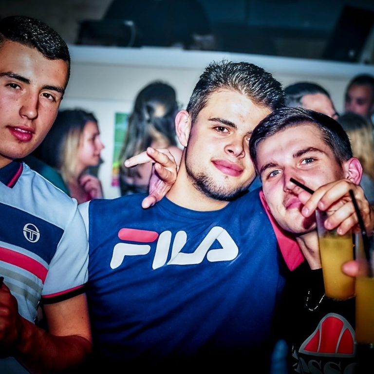 Three young men pose together in a lively social setting, holding drinks and smiling.
