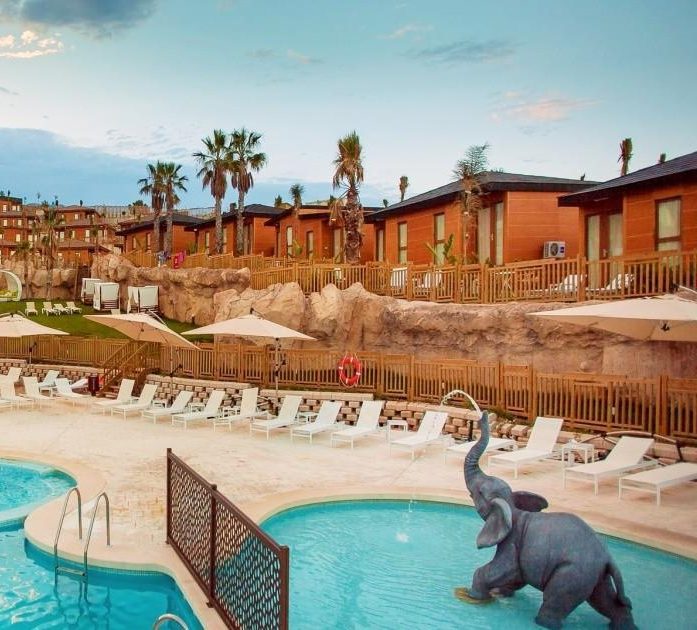 Magic Natura Animal, Waterpark & Polynesian Lodge Resort Modern hotel with a pool in the foreground and palm trees, under a cloudy sky.