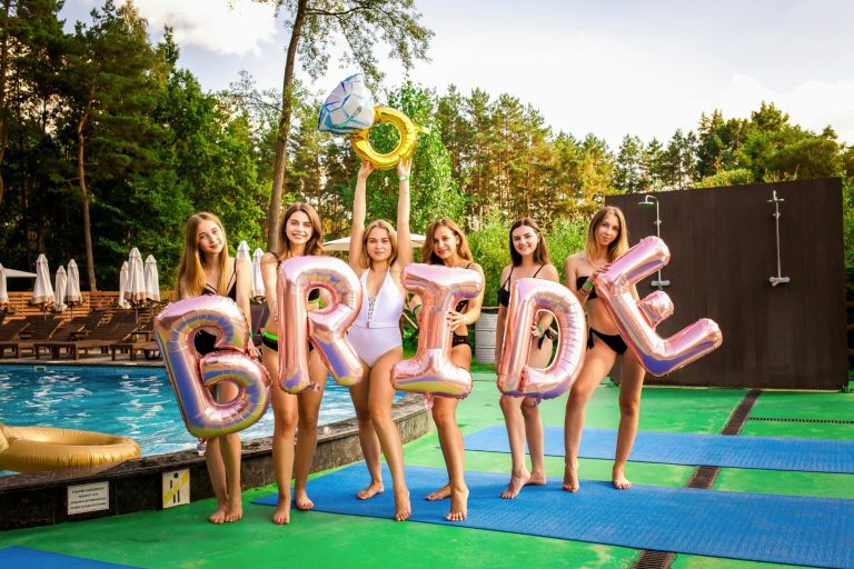 Group of smiling women in swimsuits holding "BRIDE" balloons by a poolside.