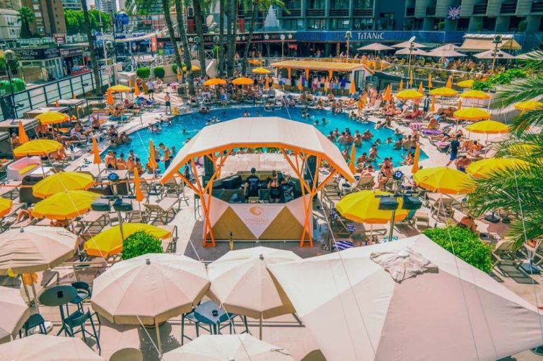 A vibrant pool area with umbrellas and sun loungers, busy with people enjoying the sun.
