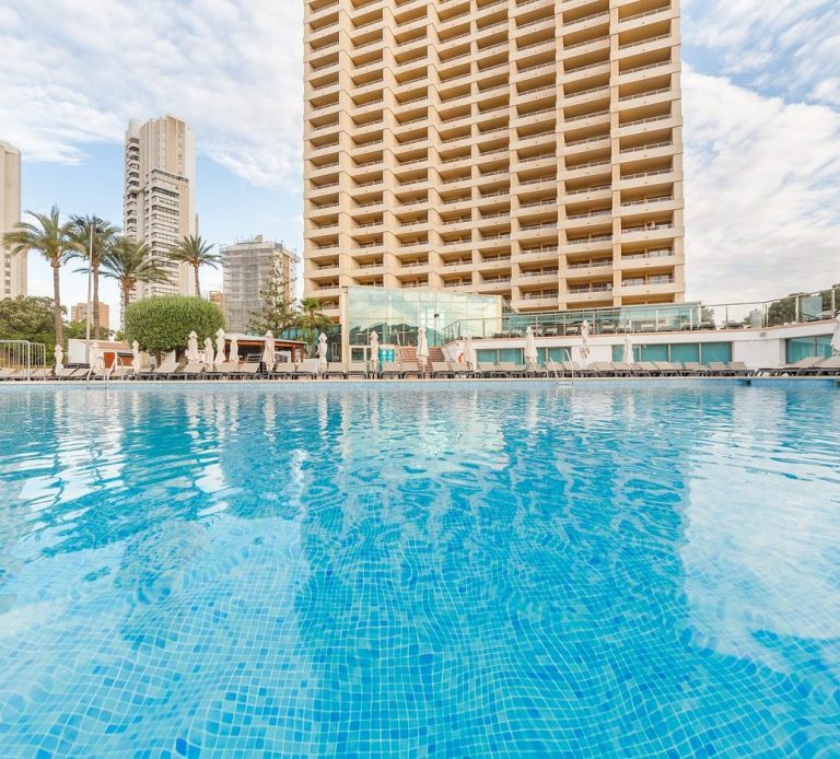 Sandos Benidorm Suites Modern hotel with a pool in the foreground and palm trees, under a cloudy sky.