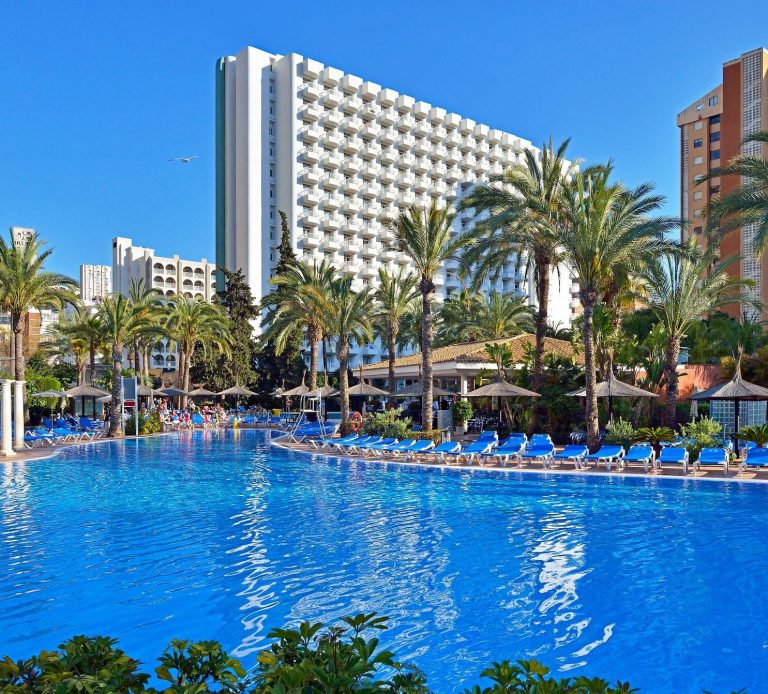Sol Pelícanos Ocas Luxury hotel pool surrounded by palm trees and sun loungers under a clear blue sky.