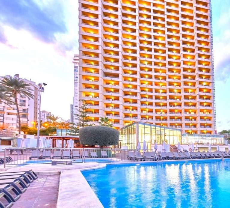 Sandos Benidorm Suites Modern hotel with a pool in the foreground and palm trees, under a cloudy sky.