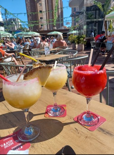 Three vibrant cocktails on a table in a lively outdoor dining area.