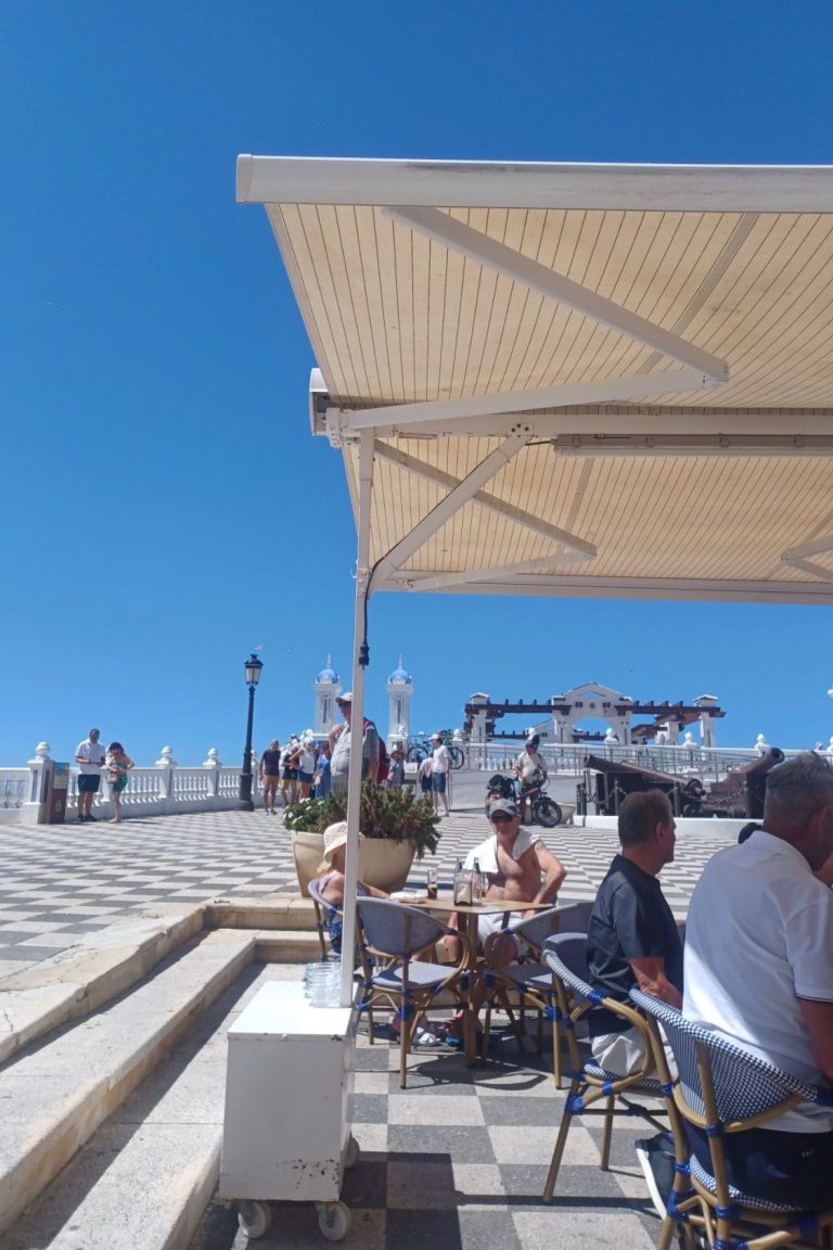 Outdoor café scene with people seated under a sunshade on a sunny day.