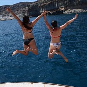 Two women jumping into blue water from a boat, holding hands, enjoying a sunny day.