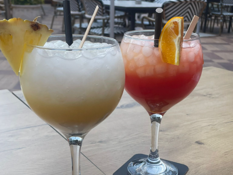 Two cocktails in glasses, one with a pineapple wedge and the other with an orange slice.